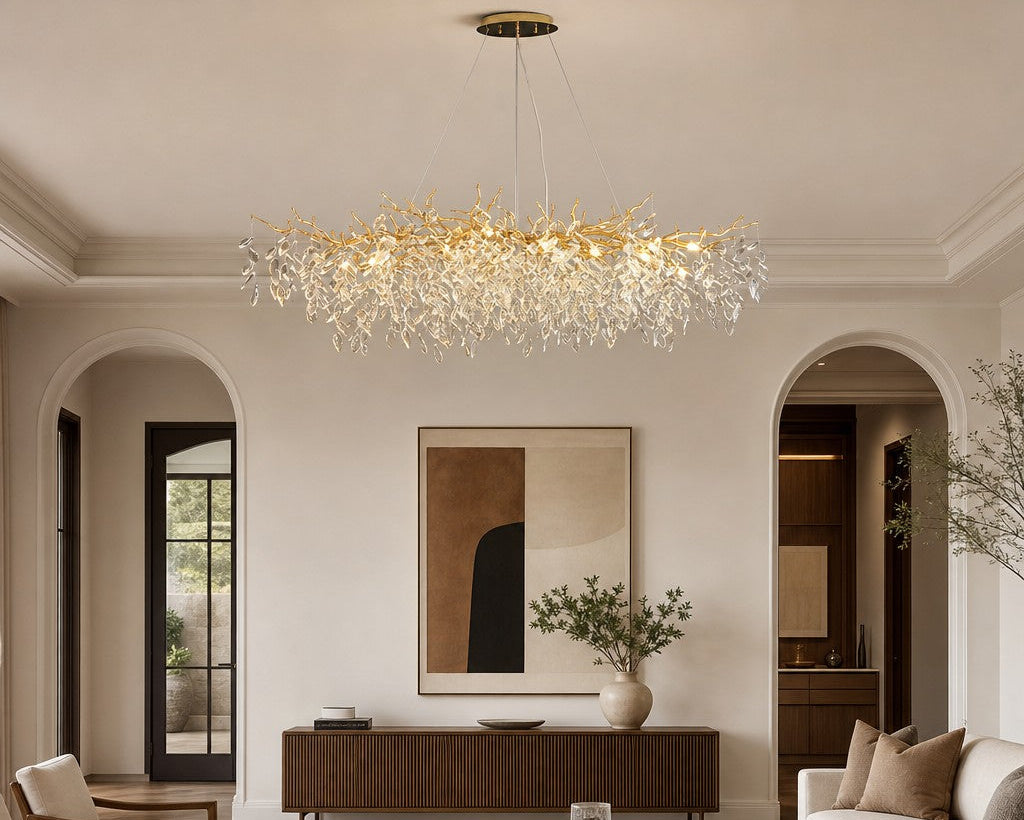A modern crystal chandelier with a gold-toned branch-like frame hangs in a bright, elegant living room with cream walls, arched doorways, a sofa, and a dark wood coffee table.